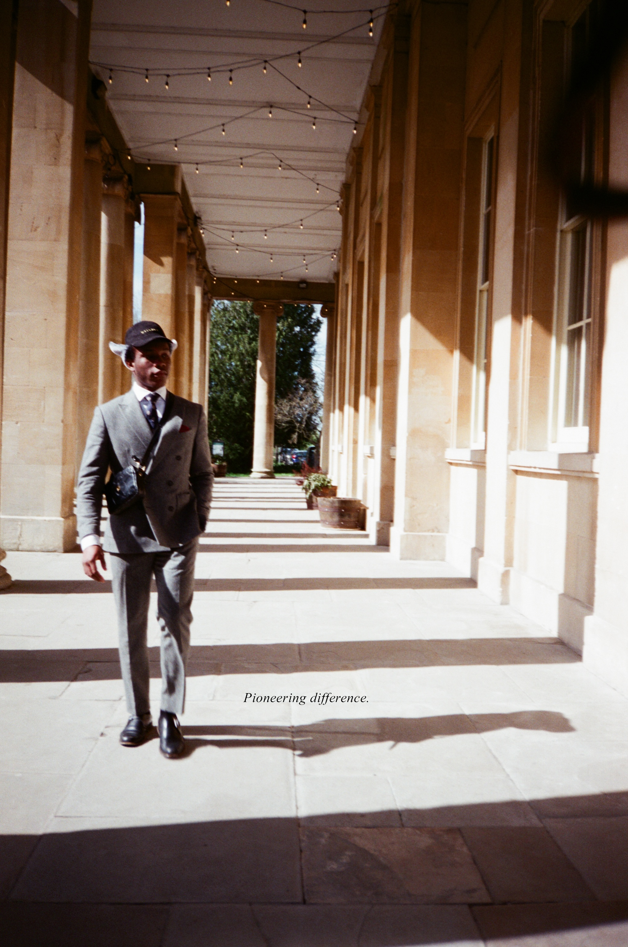 Model in a grey suit walking down a colonnade wearing the Herald trapper cap, the Acolyte cross body bag, vintage style